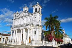 A grand white church with two bell towers stands tall under a blue sky, framed by palm trees and vibrant flowers along the surrounding walls.