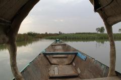A forward-facing view from a weathered wooden boat with a woven canopy, gliding through calm waters toward a distant shoreline.