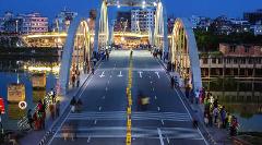 This is a wide shot of a bridge at dusk, with many people walking across it and city buildings in the background.