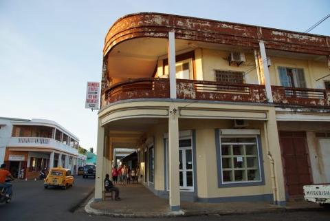 A street scene in Diego Suarez (Antsiranana) featuring an old building with peeling paint, a "Bed and Breakfast" sign, people walking on the sidewalk, and a yellow taxi on the street.