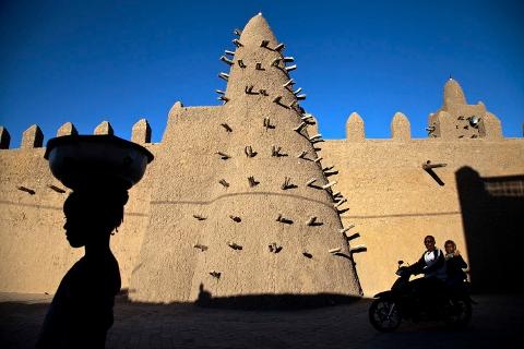 A silhouette of a woman carrying a bowl on her head and a couple on a motorcycle stand in front of the Great Mosque of Djenné in Mali.