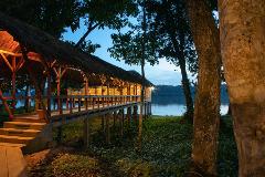 A wooden structure with a thatched roof extends over a body of water, illuminated by warm lights, with trees in the foreground against a dusk sky.