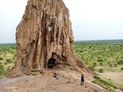 A towering rock formation with a small cave at its base stands in a vast, green, savanna-like landscape with scattered trees.