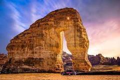 A massive natural sandstone arch dominates a desert landscape under a vibrant sky, with a vehicle parked at its base for scale.