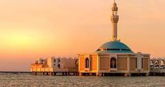 A mosque with a turquoise dome and a minaret stands on stilts in the water, bathed in the warm light of sunset.