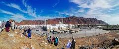 A scenic view showcases a large white mosque surrounded by a desert landscape, with mountains in the background and people traversing a rocky hill in the foreground, under a bright blue sky.