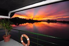 Ein atemberaubender Sonnenuntergang, der sich in einem Fluss spiegelt, vom Deck eines Schiffes aus betrachtet, mit einer Pflanze und einem Rettungsring im Vordergrund.