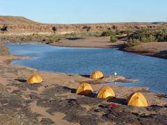 Barbecue and Camping by Niger River and Sand Dunes Five tents are pitched near a body of water, with sand dunes and cliffs in the background.