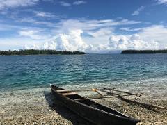 A traditional outrigger canoe rests on a pebble beach with crystal-clear water, under a sky filled with puffy clouds, and islands in the background.