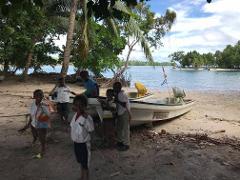 A group of children pose near two boats on a sandy beach with trees and water in the background.