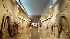 A museum hall showcases ancient sculptures of winged bulls, illuminated by overhead lighting, along a glossy floor leading to a distant exhibit.