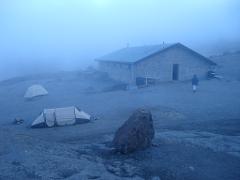 A misty mountain scene shows a stone building, two tents, a large rock, and a person walking towards the building.