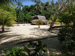 Muschu Island Day Tour From Wewak A serene tropical beach scene with a traditional thatched-roof hut nestled among palm trees and lush greenery on Muschu Island.
