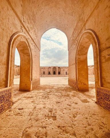 An architectural shot of an old courtyard with arched openings in warm, earthy tones, leading to a distant building under a cloudy sky.