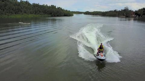 Two people ride a jet ski on a river with a boat in the distance and trees lining the shore.