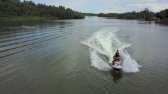 Two people ride a jet ski on a river with a boat in the distance and trees lining the shore.