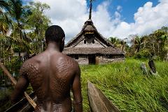 A man with patterned scarification on his back stands in a canoe, facing a traditional tribal house with a thatched roof in a lush, tropical environment.