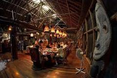 Here's a description of the image based on your prompt:A cozy, rustic interior of Karawari Lodge features wooden floors, exposed beams, tribal art, and a long dining table with guests, illuminated by hanging lamps.