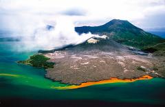 An aerial view of the Tavurvur volcano in Papua New Guinea, erupting with white smoke, surrounded by a coastline with unusual colors in the water.