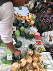 A vibrant market scene in Rabaul featuring various fruits and vegetables like watermelons, coconuts, and tomatoes, with locals selling and shopping.