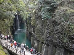 A scenic view of Takachiho Gorge with a waterfall, a turquoise river, and tourists walking along a wooden pathway.
