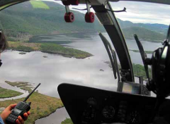 A view from a helicopter cockpit shows a lake surrounded by green hills and marshland, with a person holding a two-way radio.