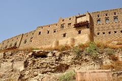 A historic brick building stands atop a crumbling hill under a clear blue sky, showcasing a blend of sturdy architecture and natural erosion.