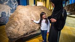 A girl points at petroglyphs on a large rock formation while standing next to a woman wearing a hijab in a museum setting.