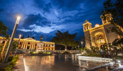 A city square at night illuminated by building lights, street lamps, and reflective wet pavement under a dramatic sky.