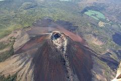 An aerial view captures the stark beauty of a volcano in Nicaragua, with its rugged slopes, verdant crater, and surrounding lush landscape dotted with a lake, possibly taken during a helicopter tour.