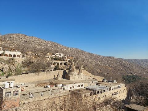 A serene landscape features tiered, tan-colored buildings cascading down a hillside, dominated by two pointed structures, under a clear blue sky.