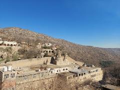 A serene landscape features tiered, tan-colored buildings cascading down a hillside, dominated by two pointed structures, under a clear blue sky.
