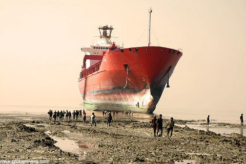 A large red ship is beached on a muddy shore, with a group of people standing and walking around it.