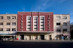Here's a one-sentence description of the image:The Cinema Impero, an art deco style building with "CINEMA" and "IMPERO" inscribed vertically on either side of its facade, stands in an urban setting under a clear blue sky.