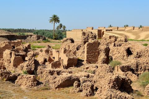 The image shows the ruins of an ancient brick structure, likely in the Middle East, with a restored brick wall and palm trees in the background under a clear blue sky.