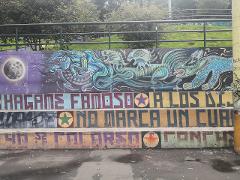 A colorful urban art mural depicting mythical creatures and bold Spanish text on concrete steps, against a backdrop of trees and a metal railing.