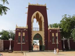 The image showcases the Gidan Rumfa, the Emir's Palace in Kano, with its striking red and beige entrance gate under a clear sky.