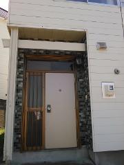 A beige door with a metal grate and a white panel is seen in front of a building with white horizontal siding and a decorative brick-patterned column.