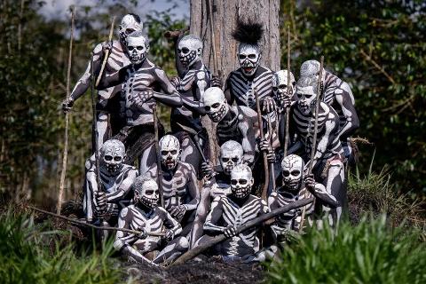 A group of men from the Skeleton Tribe are gathered together, wearing intricate white skeleton paint on their black bodies, holding sticks, and posing in front of a tree in the highlands of Papua New Guinea.