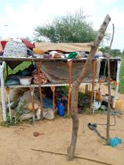 A makeshift two-tier bed frame stands outdoors, laden with blankets, baskets, and various items, with a leaning tree trunk propped against the front.