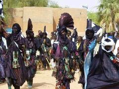 A group of men in traditional attire, including face coverings and ornate hats, participate in the Bianou Festival in Niger, with buildings and a tree visible in the background.