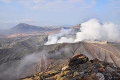 A scenic view of Mount Aso, an active volcano in Japan, featuring steam rising from its caldera with a rocky foreground.
