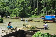 5-Day Dhaka Cultural Heritage Tour: Floating Markets & River Life Experience Men in boats transport sugarcane down a muddy river, with lush green foliage lining the banks and a blue and red larger boat moored to the side.