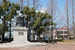 A statue of a woman holding a baby stands on a pedestal in front of trees and buildings under a blue sky. The pedestal has the inscription "1945 8.9 11:02".