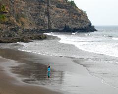 A woman in a blue skirt walks along a wet, dark sand beach with waves crashing nearby, set against a backdrop of a high, rocky cliff.