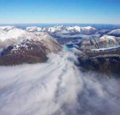 Here's a description of the image:An aerial view shows a valley filled with a thick layer of fog or low clouds, surrounded by rugged mountains, some with snow-capped peaks, under a clear blue sky.
