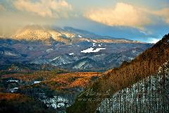 A scenic view of a mountainous landscape with patches of snow, forests, and clouds with golden sunlight hitting the mountaintop.