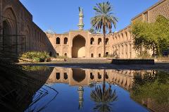 A serene courtyard with tan buildings and a distinctive tower is reflected in a pool, creating a symmetrical and visually striking scene under a clear blue sky.