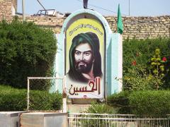 A poster of a man with a beard and green turban is framed by a light blue arch, surrounded by green bushes, a brick wall, and a white fence.