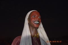 A person with a painted red face and black lips, wearing a white head covering and a colorful necklace, smiles into the darkness.